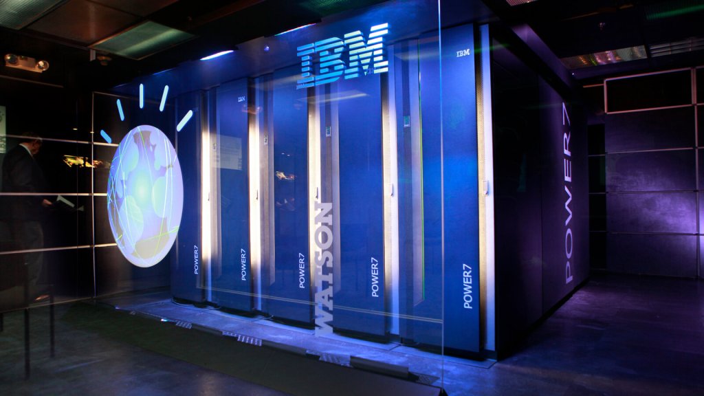 A sleek, futuristic image of IBM's Watson, a powerful AI-driven supercomputer system, housed in large, vertical server racks labeled "POWER7". The glowing IBM logo and Watson’s signature globe icon are prominently displayed on the glass panel, highlighting advanced data processing and artificial intelligence capabilities. The background features a high-tech, dimly lit room, emphasizing the modernity and cutting-edge nature of this technology.